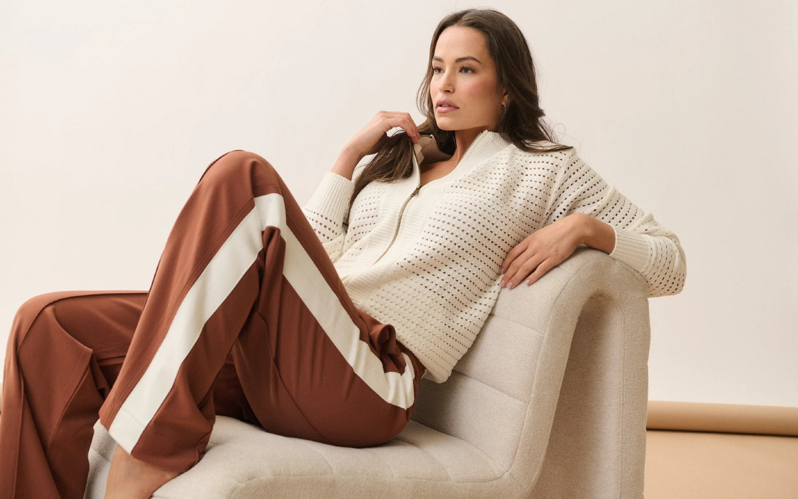 Woman sitting on a white chair wearing a beige knitted cardigan and brown pants from Cartel & Willow